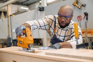 Services black african male carpenter joiner using jig saw wood panel cutter to cut making furniture panel in workshop photo